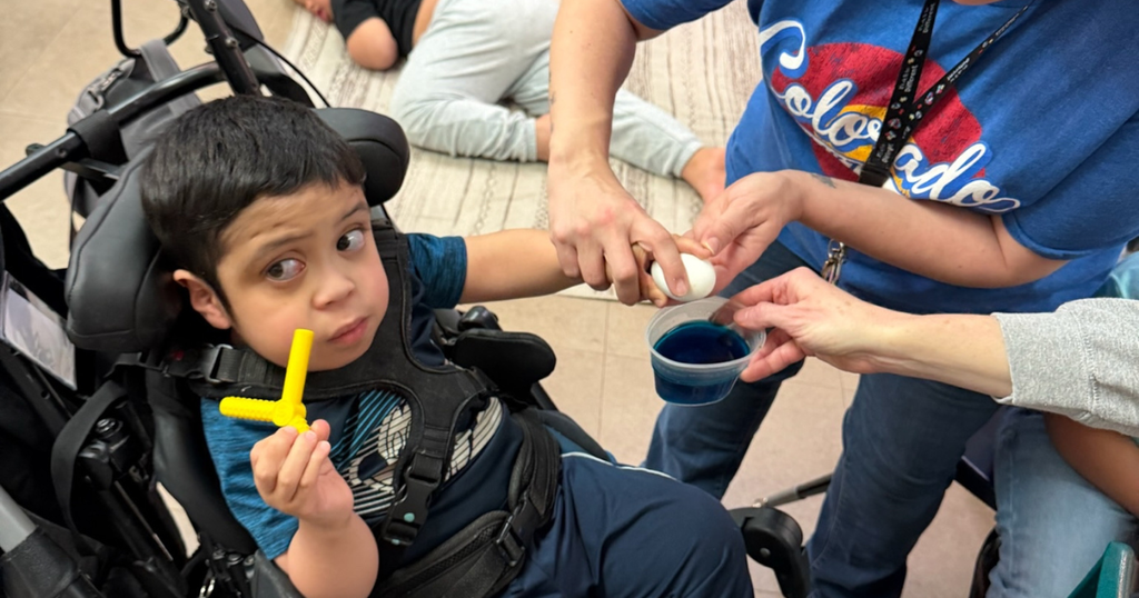 A student seated in a supportive chair holds a yellow tool while adults assist him in dipping an egg into blue dye.