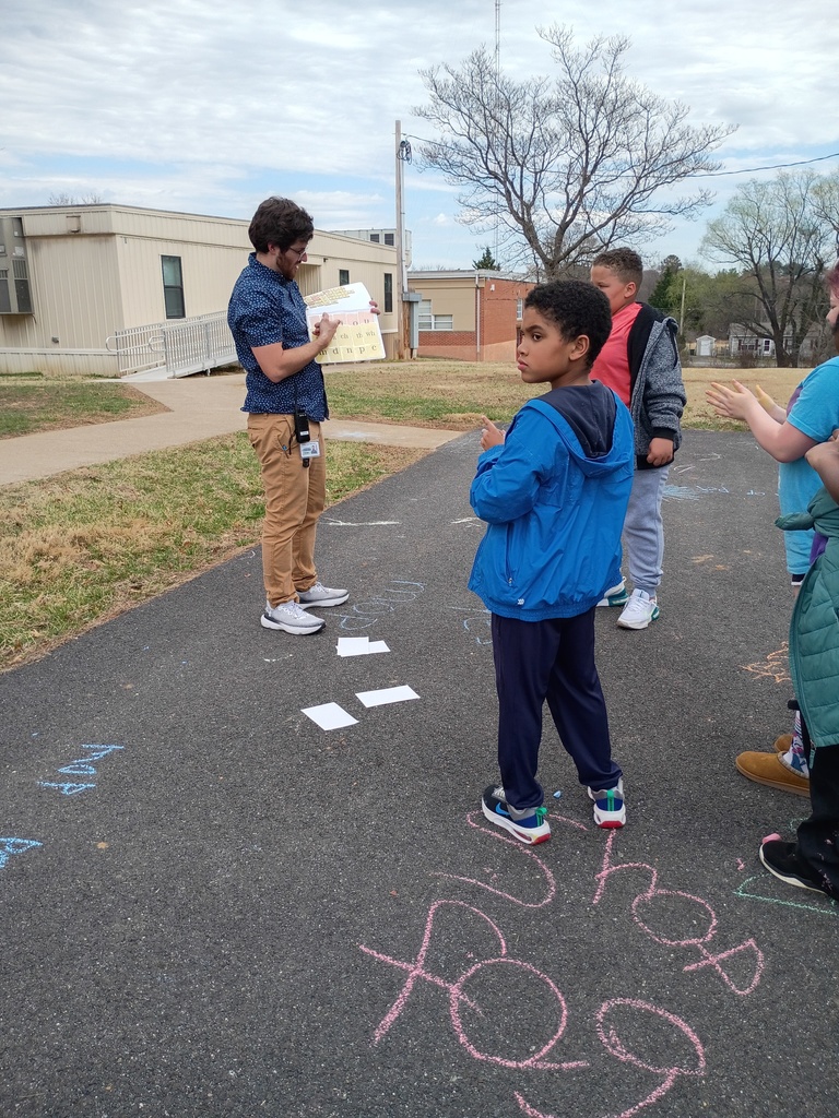 A teacher stands on an asphalt path holding a phonics board for a group of students. The focus is on the back of a boy in a blue jacket who has his hands on his head while looking at the board. Several white cards are placed on the ground in front of the teacher, and various chalk words are drawn on the pavement nearby.