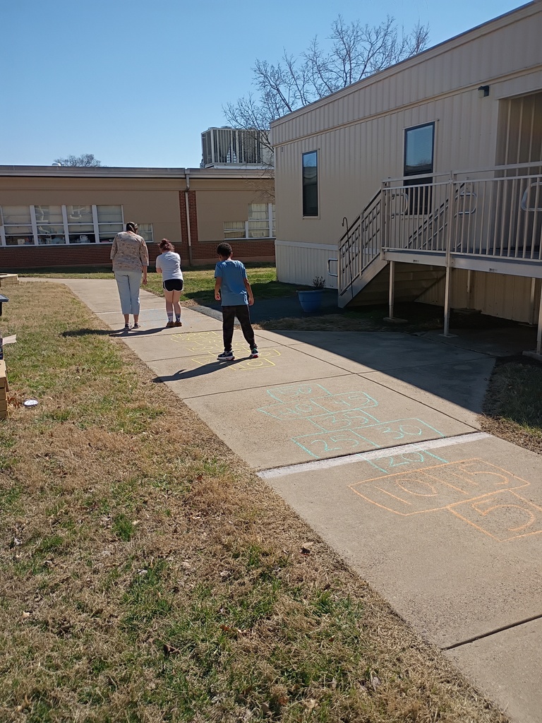 A wide outdoor shot of a concrete walkway leading toward school buildings. Three people—an adult and two children—are walking away from the camera. In the foreground, colorful chalk hopscotch-style grids are drawn on the pavement, containing numbers like 5, 10, 15, and up to 50, suggesting a math-based jumping game.