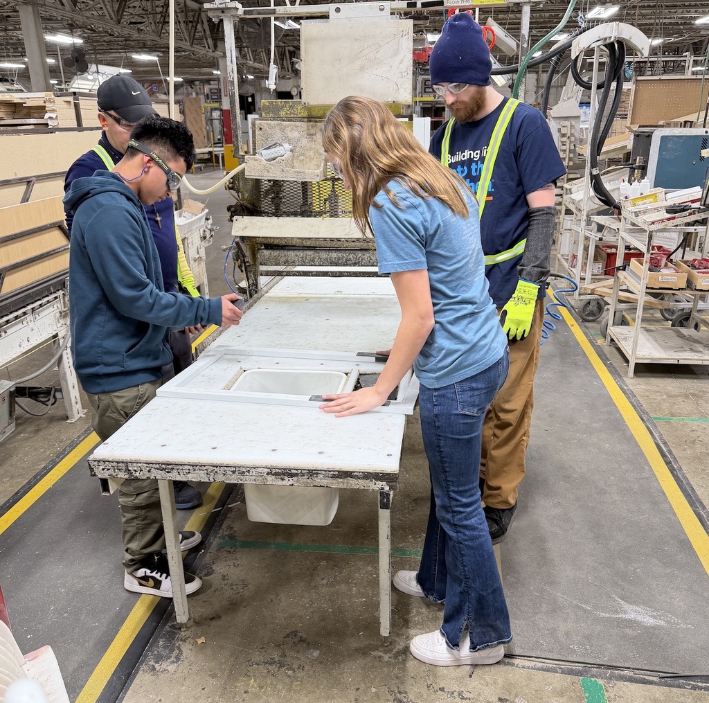 Three students and an instructor work together around a table in an industrial workshop. They are positioning and securing a rectangular sink or fixture into a countertop while using tools and wearing safety gear such as gloves and protective eyewear. The space includes large machinery, hoses, and marked floor areas, indicating a hands-on manufacturing or technical training environment.