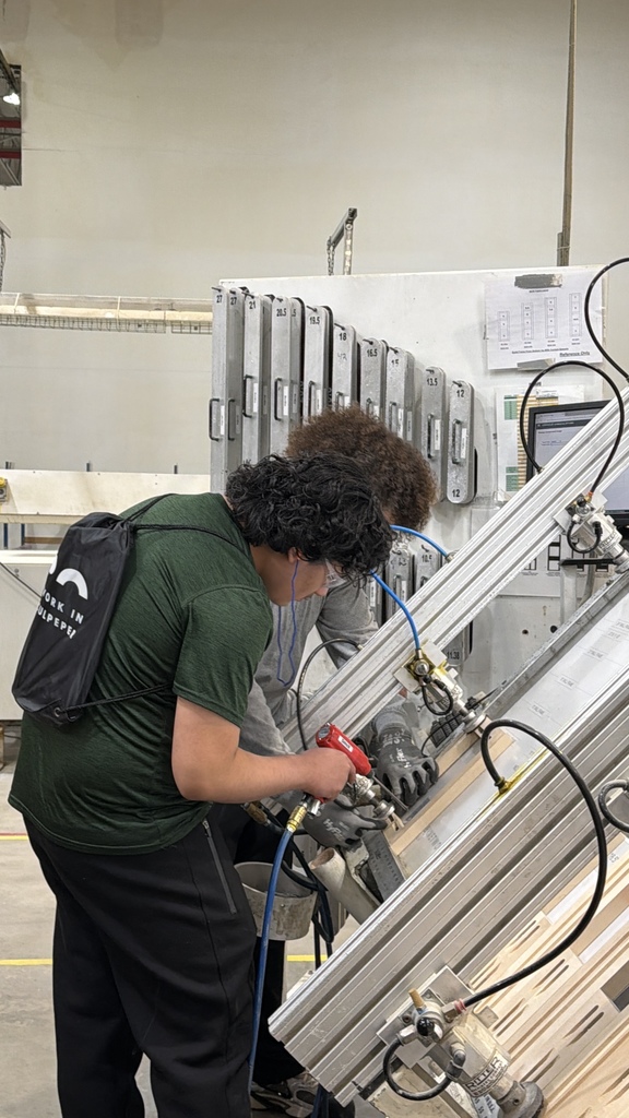 Two students work together using a pneumatic tool at an angled workstation in a manufacturing or technical lab setting. One student operates the tool while the other assists, both wearing safety gloves. The area includes industrial equipment, hoses, and labeled panels, indicating hands-on technical training.
