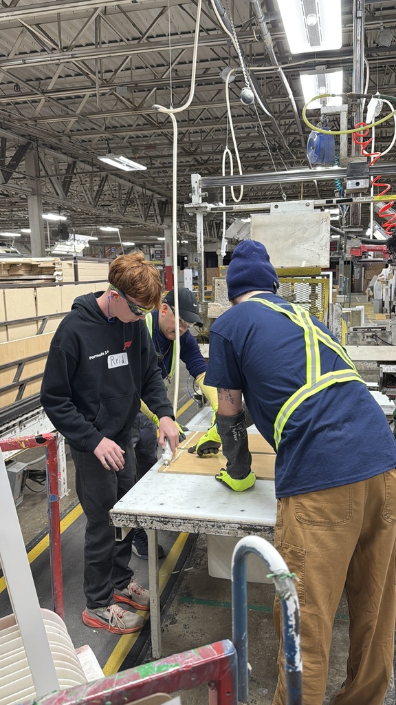Three people work together at a table in an industrial workshop or manufacturing facility. One person applies adhesive or sealant to a flat board while the others assist, wearing safety gear including gloves and protective eyewear. The space includes machinery, overhead lighting, and materials stacked nearby, suggesting a hands-on technical or vocational activity.