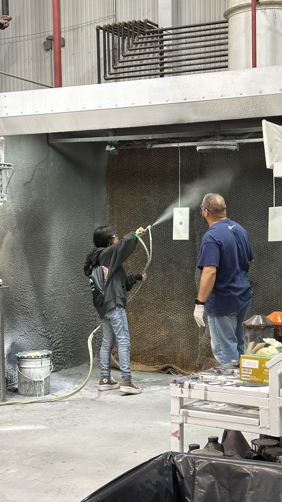 A student uses a spray gun to coat a hanging panel inside an industrial spray booth while an instructor stands nearby supervising. The student wears casual clothing and holds a hose connected to the sprayer, directing a fine mist toward the surface. The workshop environment includes ventilation panels, pipes, and paint supplies, indicating a hands-on technical training activity.