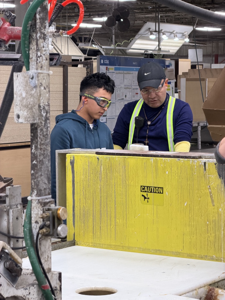 A student wearing safety goggles stands beside an instructor in an industrial workshop as they operate or examine a large machine with a yellow safety guard labeled “Caution.” The instructor gestures while explaining the process, and both focus on the task. The space includes manufacturing equipment, overhead lighting, and materials, indicating hands-on technical training.