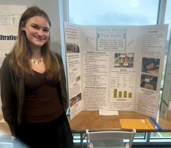 Student poses beside a project titled “Terra Prima (First Earth),” with research, photos, and charts about early Earth conditions and experiments.