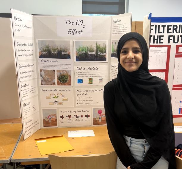 Student stands in front of a display titled “The CO₂ Effect,” showing plant growth experiments and images comparing results under different conditions.