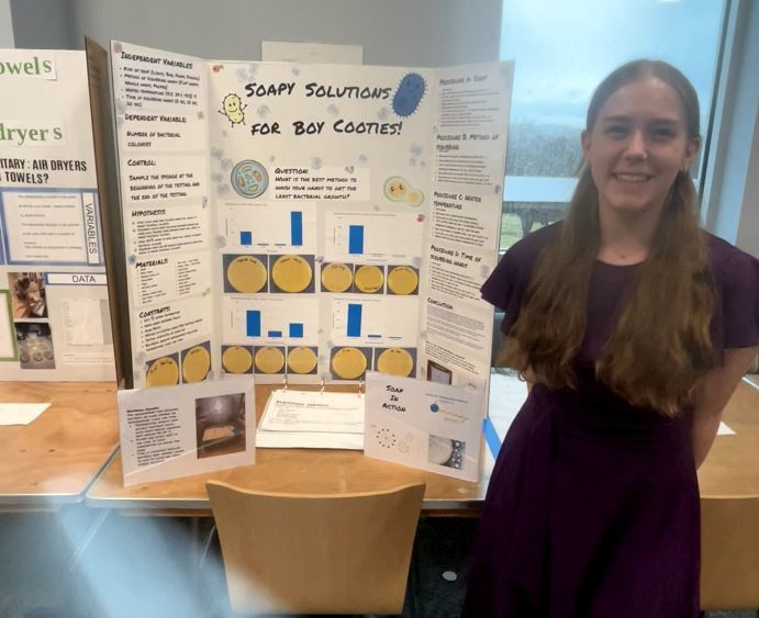 Student stands next to a science fair project titled “Soapy Solutions for Boy Cooties,” displaying petri dish results and bar graphs showing bacteria growth.