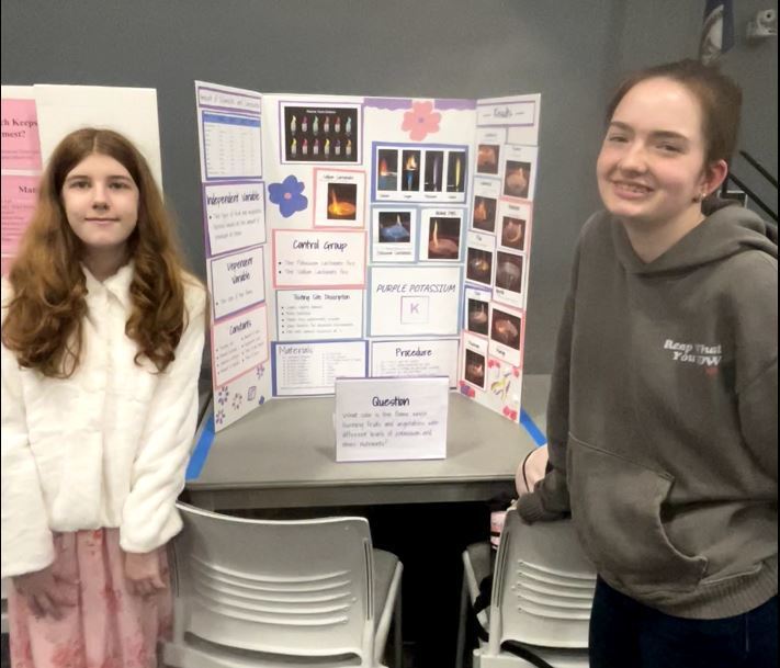 Two students stand beside a science fair display featuring flame test experiments with potassium, including labeled photos, procedures, and results.