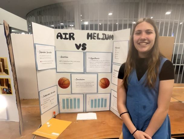 Student stands smiling beside a science fair display titled “Air vs Helium,” featuring charts, graphs, and basketball illustrations comparing the two gases.