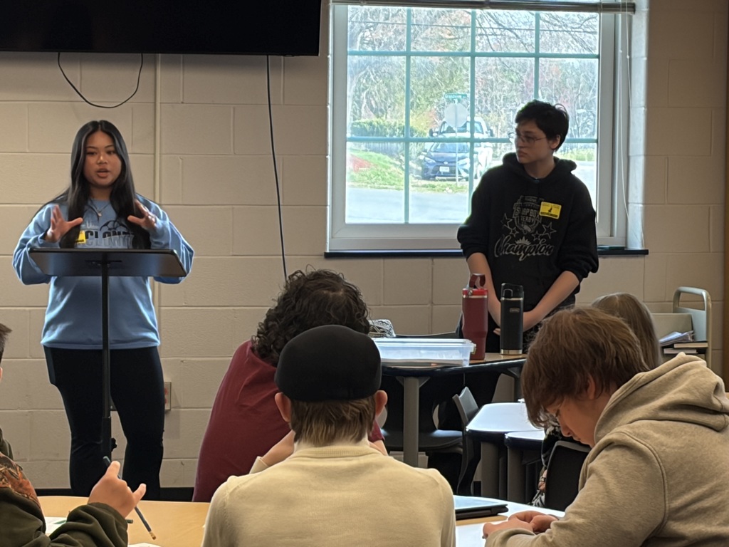 A student stands at a podium in a classroom presenting to peers, gesturing while speaking. Another student stands nearby at a table with materials, while several classmates sit at desks listening and taking notes. A window and classroom wall are visible in the background.