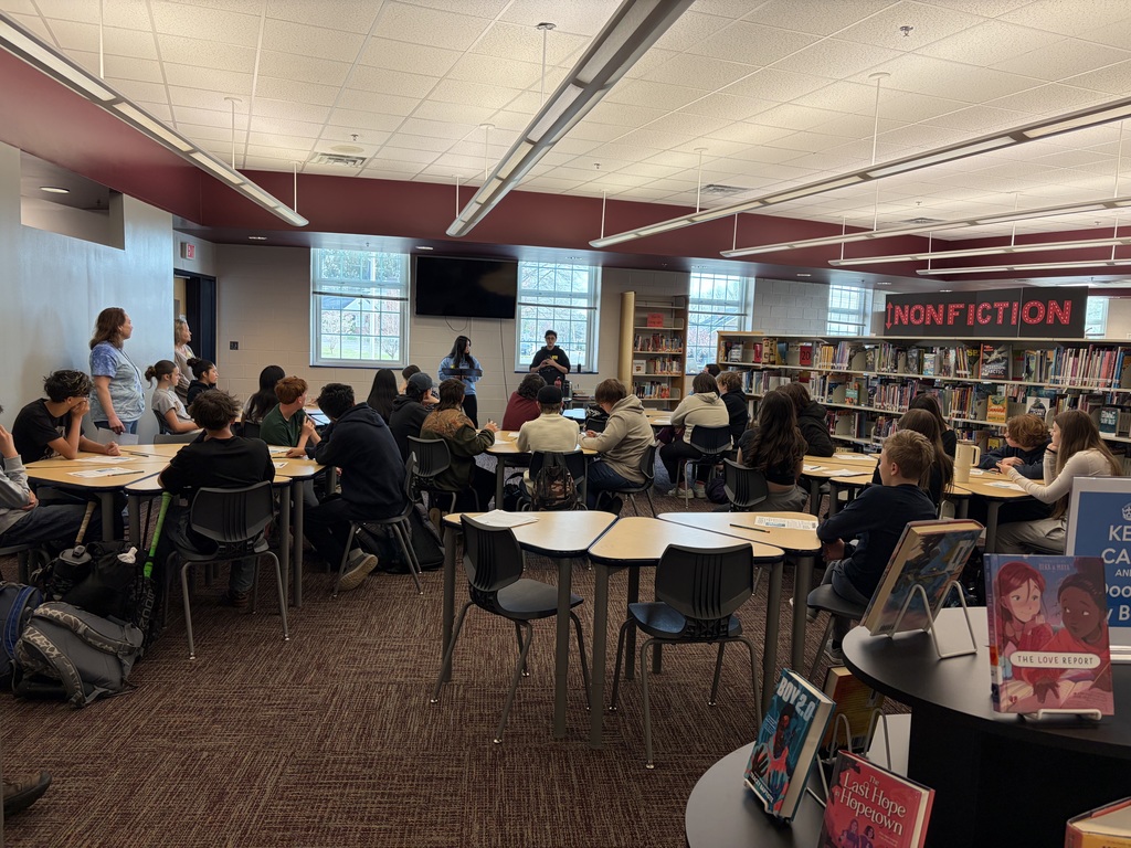 A large group of students sits at tables in a school library during a class activity or presentation. Two students present at the front of the room while others listen. Bookshelves labeled “Nonfiction” and displays of books are visible, along with teachers standing along the side observing.