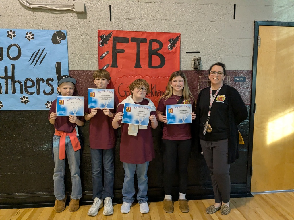 Five students stand in a school hallway holding “Excellence” certificates alongside a staff member. The students wear matching maroon shirts and smile while posing in front of colorful posters, including one that reads “FTB” with hawk imagery. The group appears to be recognized for an achievement or participation in a school program.