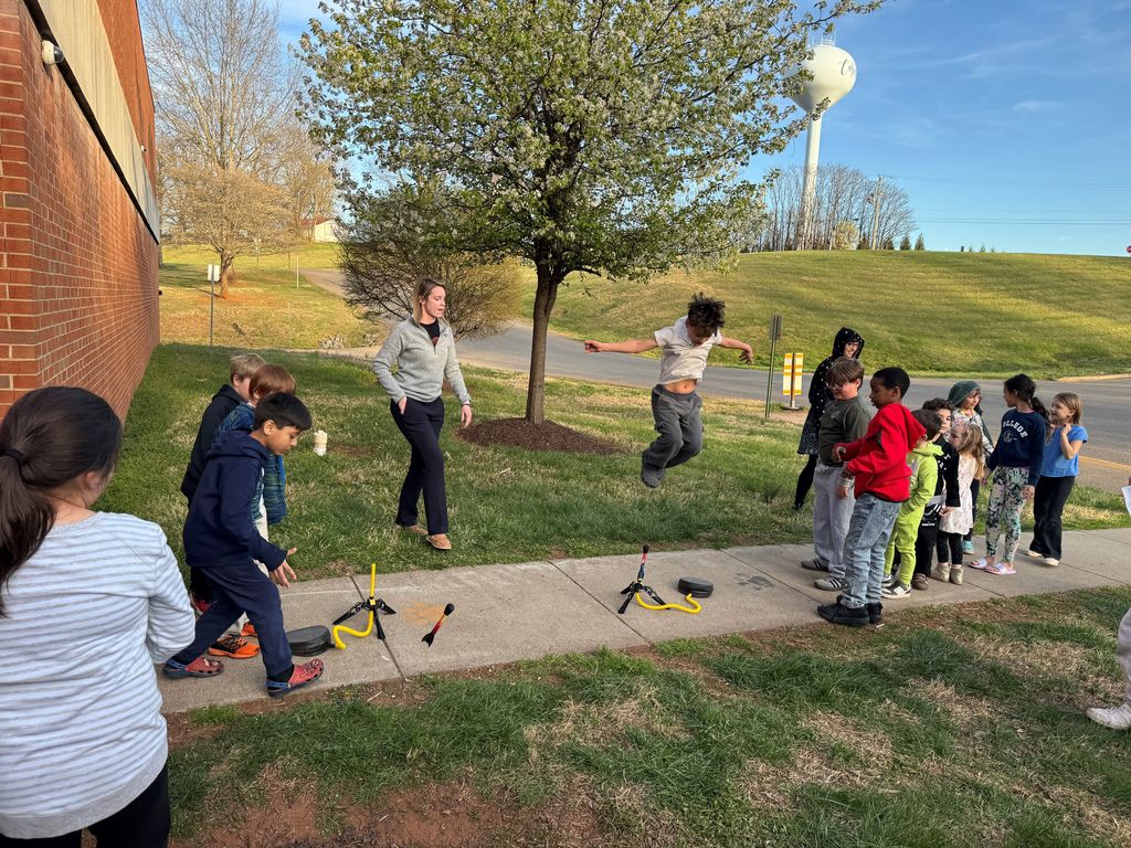 Students outside take turns launching air rockets while others line up and watch, with a teacher supervising nearby.