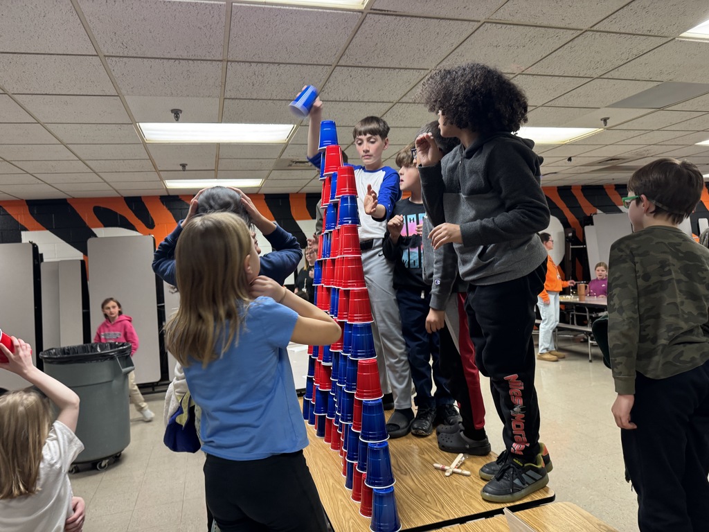 Students stack red and blue plastic cups into a tall tower while others watch and assist in a cafeteria setting.