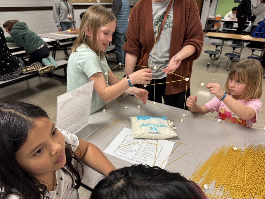 Students and an adult build 3D structures using spaghetti and mini marshmallows at a table covered with materials.
