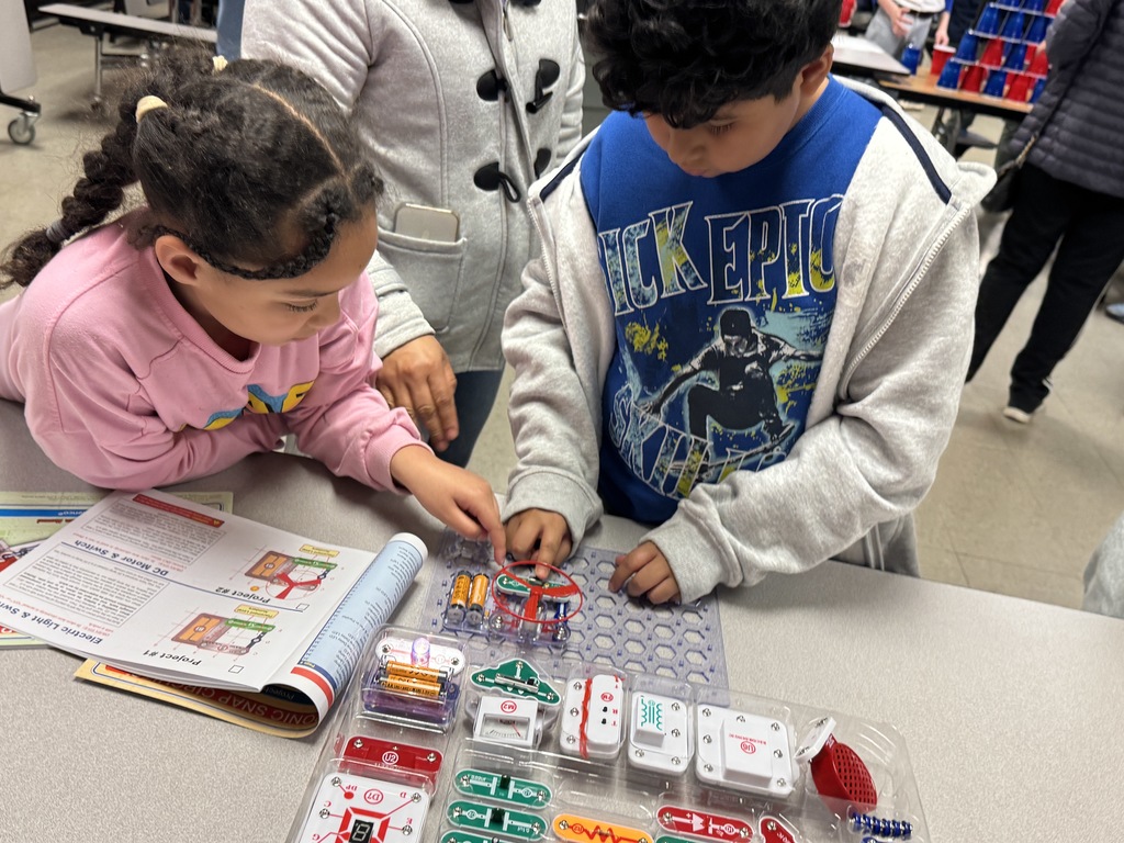 Two students work together on a Snap Circuits project, placing components onto a board while referencing instructions.