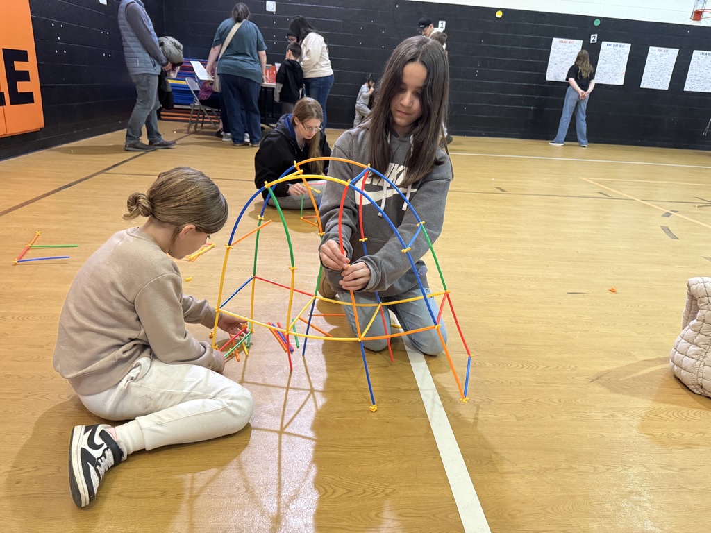 Two students assemble a colorful dome-like structure using plastic rods and connectors on a gym floor.