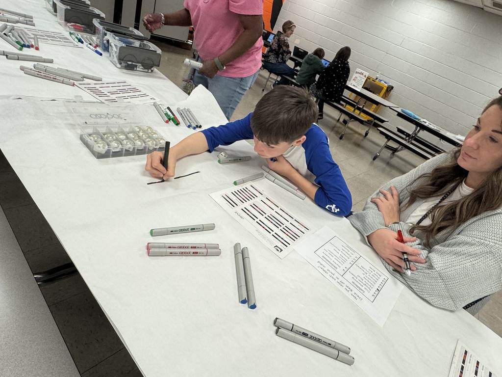 A student draws lines on paper with markers at a STEM station while an adult sits nearby; tables are covered with markers and activity sheets.