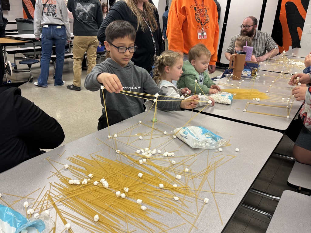 Students construct geometric structures with spaghetti and marshmallows, with materials scattered across the table.