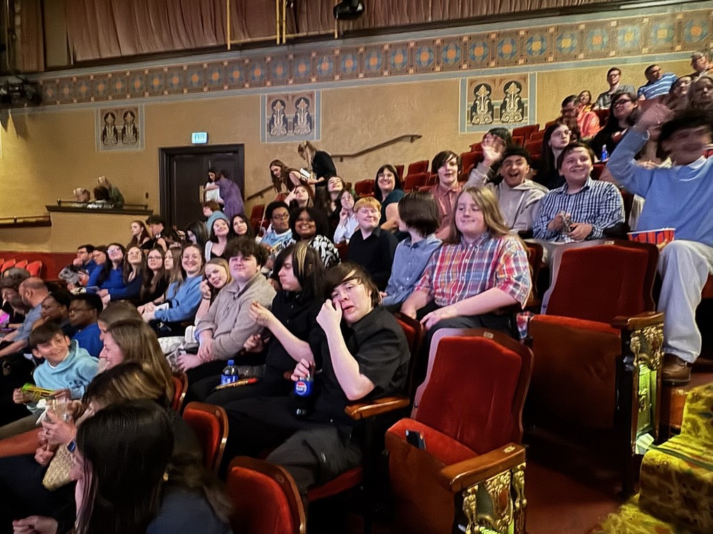 A group of students sits together in red theater seats inside an ornate auditorium, smiling, waving, and posing for a photo before a performance.