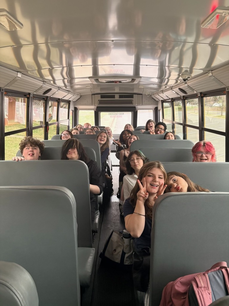 Students sit on a school bus, smiling and making peace signs while looking toward the camera during a field trip.