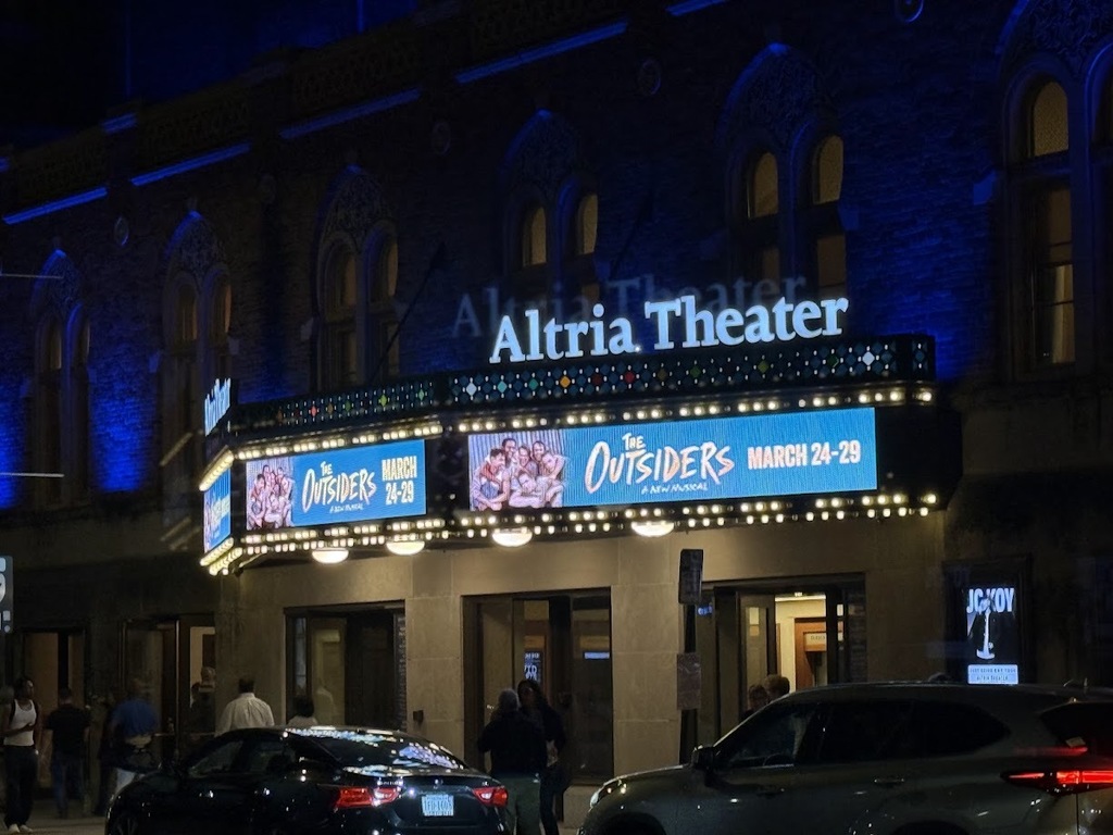 The exterior of Altria Theater at night, with a lit marquee advertising “The Outsiders” musical and people gathered outside the entrance.