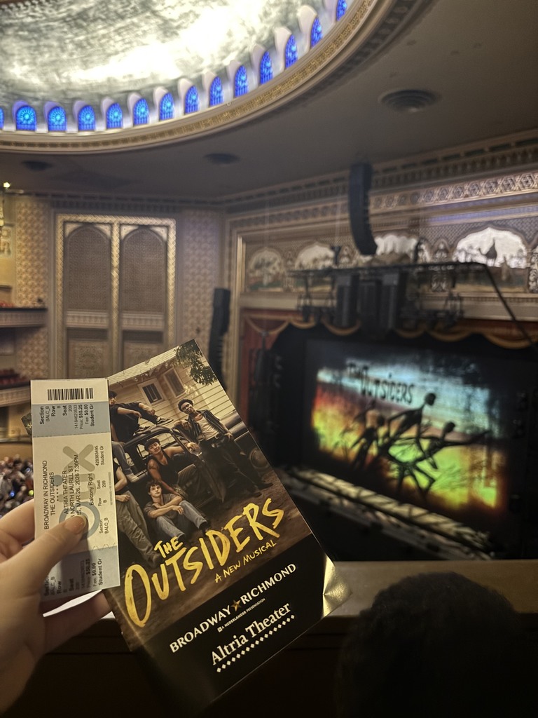 A hand holds a program and ticket for “The Outsiders: A New Musical” at Altria Theater, with the stage and set visible in the background.