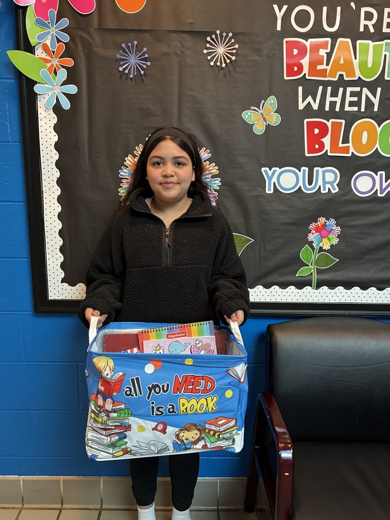 A young girl in a black fleece pullover and headband stands in front of a blue wall and a decorative bulletin board that reads "YOU'RE BEAUTIFUL WHEN YOU BLOOM." She is holding a blue fabric bin by its white handles. The bin is filled with books and colorful notebooks, with the text "all you NEED is a BOOK" printed on the front.