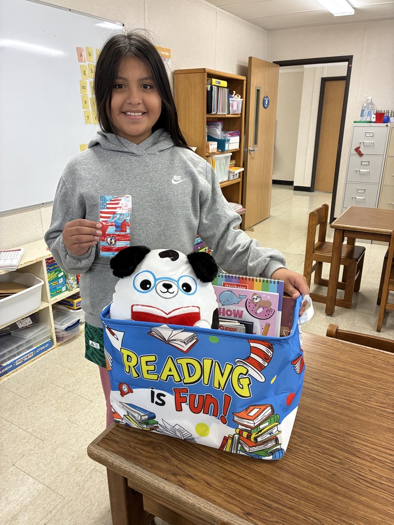 A young girl with long dark hair, wearing a grey Nike hoodie, stands in a classroom. She holds up a colorful handmade bookmark featuring an American flag design. In front of her on a wooden table is a blue fabric bin that says "READING is Fun!" filled with books and a panda plush toy wearing blue glasses.