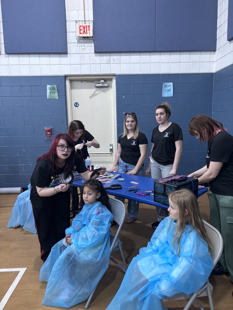 A group of older students in black "Cosmetology" t-shirts stand behind a blue table, assisting younger students. One teen with red hair is styling a young girl's hair, while the younger girl sits in a chair wearing a light blue protective gown.