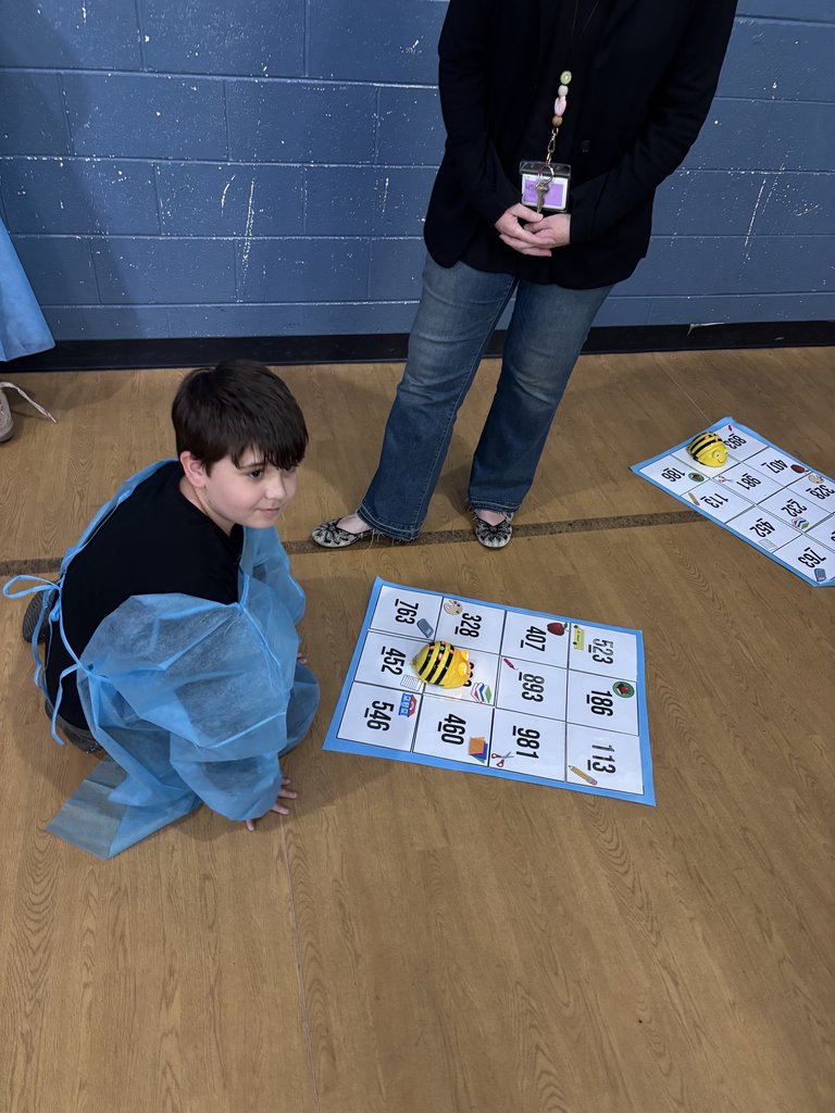 A young boy in a blue gown kneels on a wooden gym floor next to a white grid mat. On the mat, a small yellow and black toy robot (resembling a bee) sits on a square with numbers and icons.
