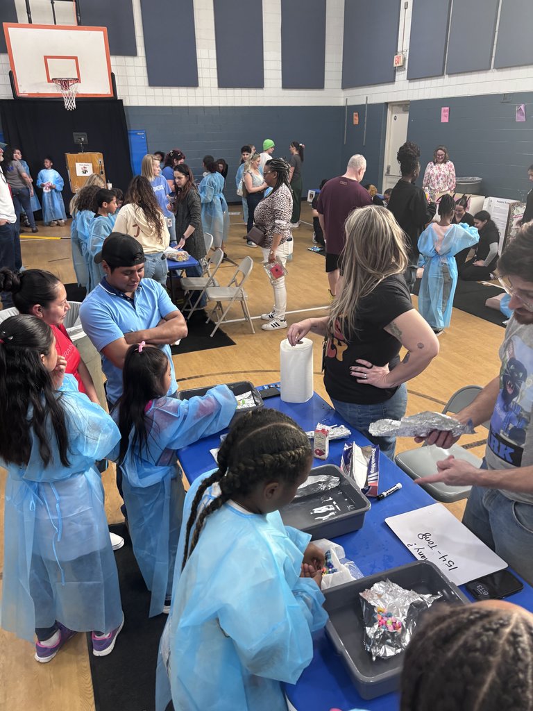 A high-angle shot of a busy school gymnasium filled with people. In the foreground, children in blue medical gowns stand at a blue table, participating in a hands-on activity involving trays and tinfoil, while adults observe.