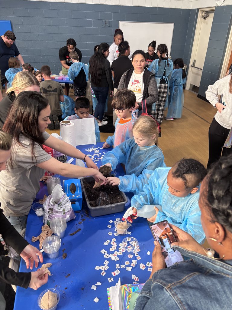 A crowded science station where a woman assists several children in blue gowns with a planting activity. They are gathered around a gray bin of soil on a blue table covered in small white stickers.