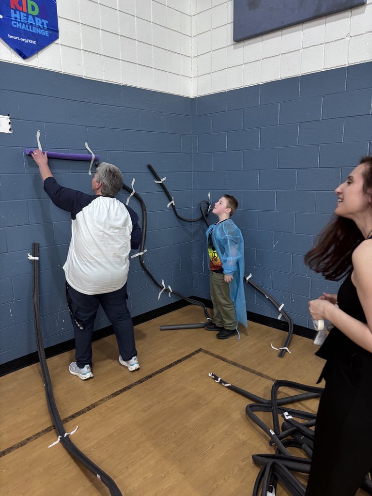 In the corner of a gym with blue brick walls, an adult and a young boy in a blue gown work together to tape long, grey foam tubes to the wall, creating a makeshift marble run or track.