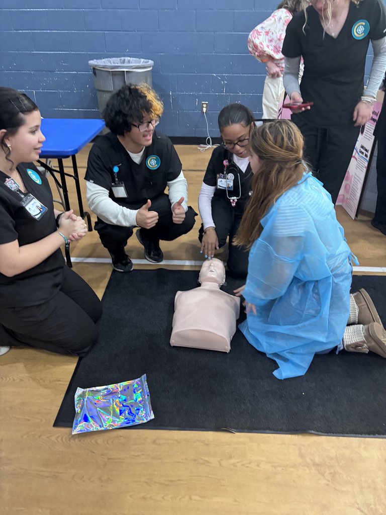 Three students in black medical scrubs kneel on a gym floor around a CPR manikin. They are instructing a younger student, who is wearing a blue medical gown, on how to perform chest compressions.
