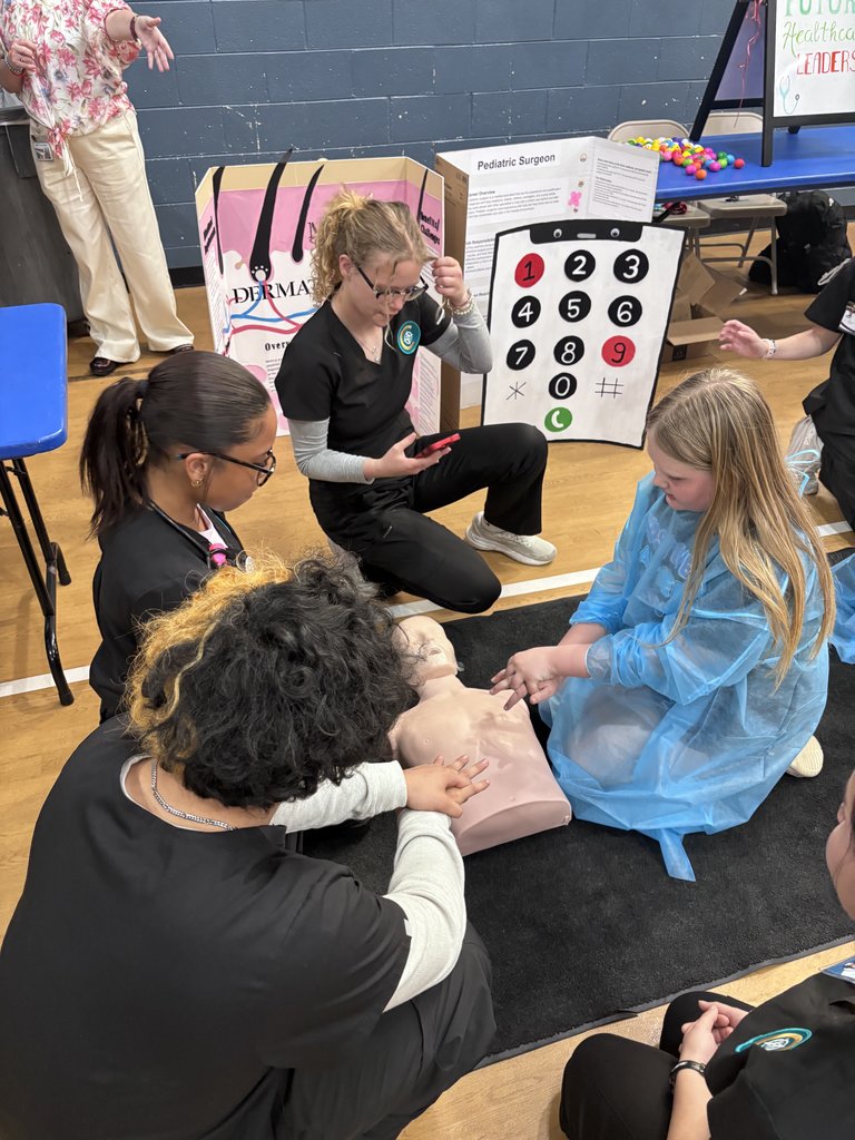 Three students in black medical scrubs kneel on a gym floor around a CPR manikin. They are instructing a younger student, who is wearing a blue medical gown, on how to perform chest compressions.