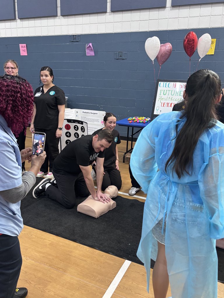 An adult male kneels on a black mat in a gym, demonstrating CPR on a manikin. Several students in scrubs and a young girl in a blue gown observe, while another person records the demonstration on a smartphone.