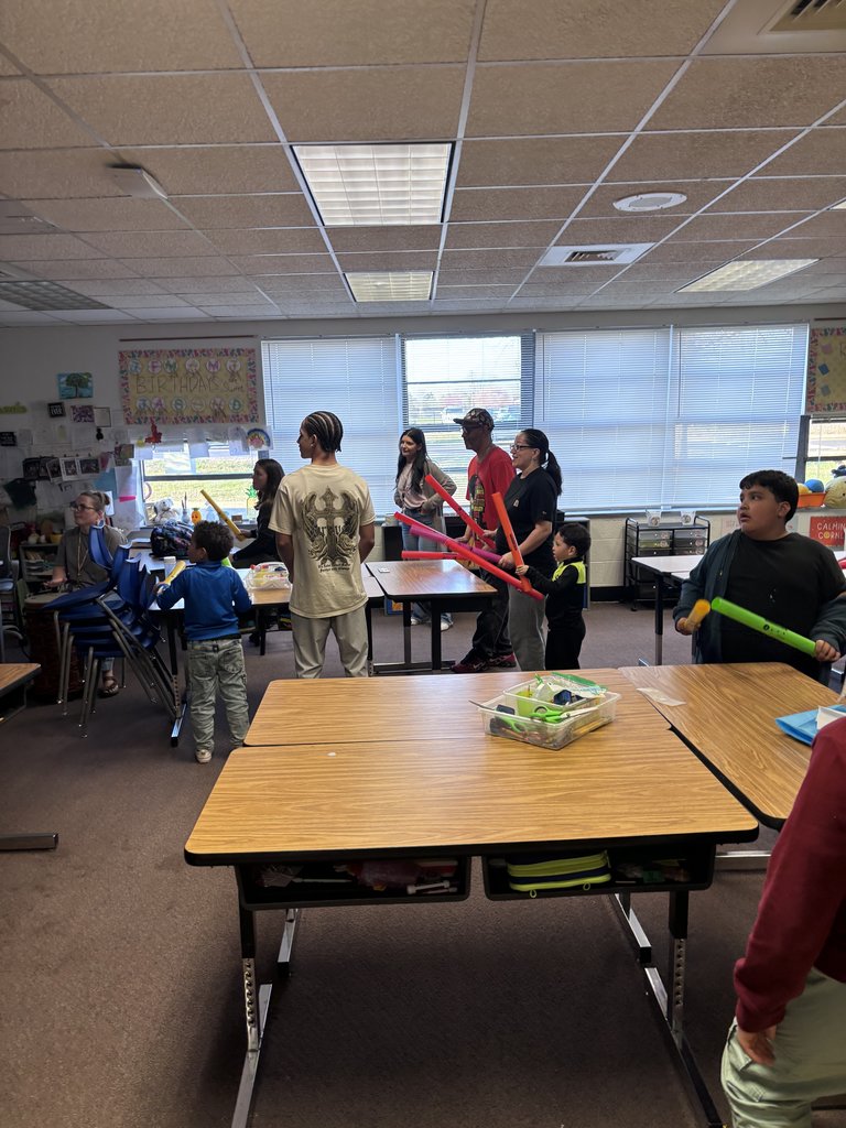 Inside a classroom, a group of students and adults stand near wooden desks. Several people are holding long, brightly colored foam "Boomwhackers" (musical tubes), appearing ready for a group activity or lesson.