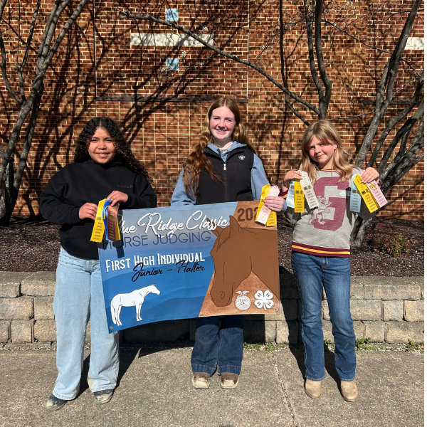 Three students stand outdoors in front of a brick wall holding a large Blue Ridge Classic Horse Judging banner and award ribbons. They are smiling and displaying their team accomplishment from the competition.