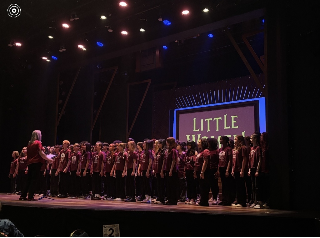 Students perform on stage in coordinated shirts while a conductor leads them from the front. Stage lights shine overhead, and a screen behind them displays “Little Women.”