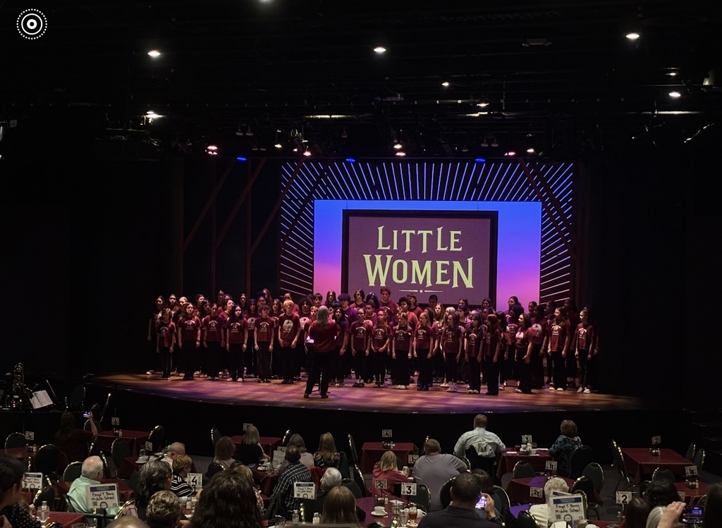 A large group of students stands on stage performing in a choir under colorful stage lighting, with a screen behind them displaying the title “Little Women.” Audience members are seated at tables in the foreground watching the performance.