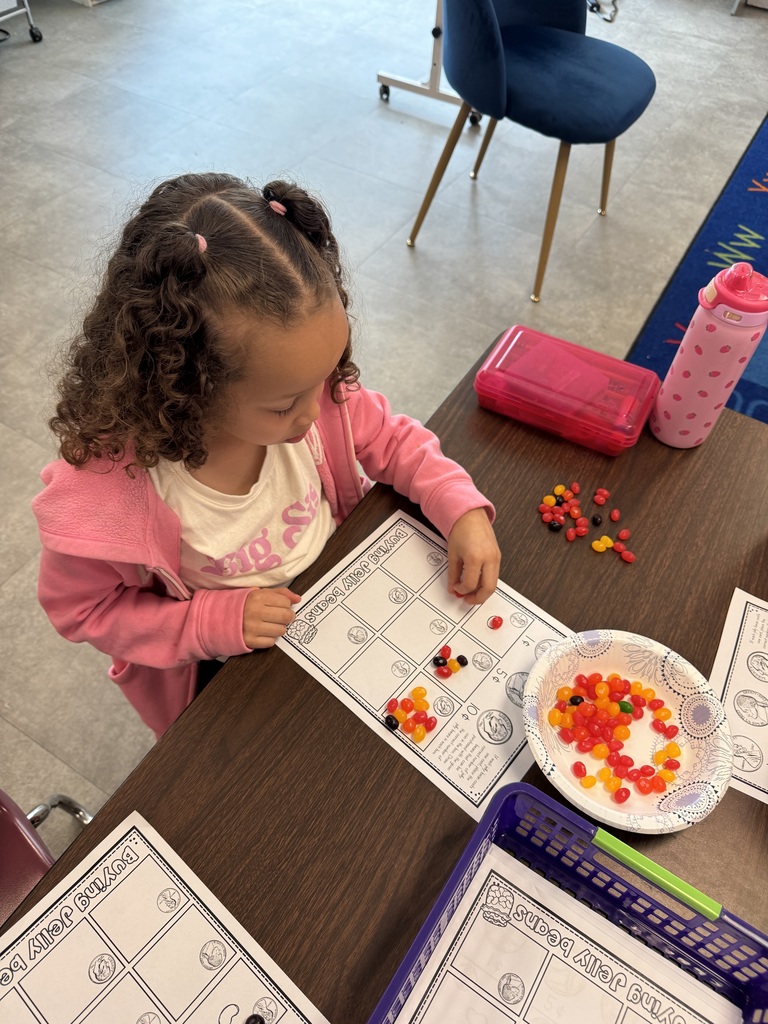 A young student in a pink jacket places jelly beans onto a counting worksheet while working independently at a desk.