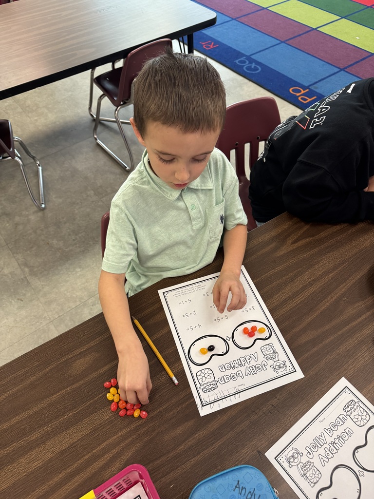 A student uses jelly beans to solve an addition problem on a worksheet, carefully counting pieces to find the answer.
