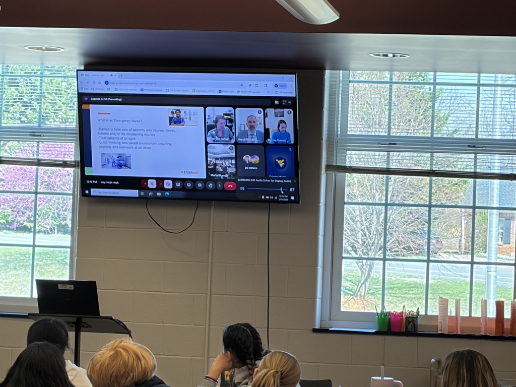 A screen at the front of a classroom displays a virtual presentation about emergency nursing, with multiple speakers visible in a video call. Students sit facing the screen, watching and listening as part of a lesson or guest presentation.