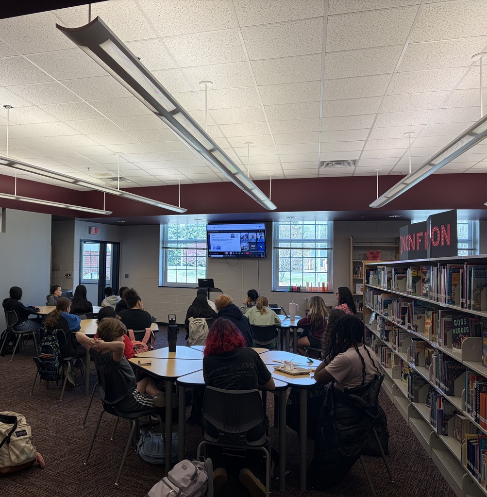 A screen at the front of a classroom displays a virtual presentation about emergency nursing, with multiple speakers visible in a video call. Students sit facing the screen, watching and listening as part of a lesson or guest presentation.