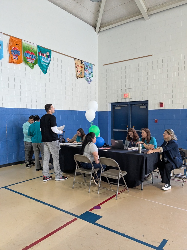 Several adults and a few children are gathered in a school gymnasium around tables covered with papers and laptops. Staff members sit and speak with individuals who are standing or seated nearby. The space has high ceilings, banners on the wall, and balloons decorating one of the tables.