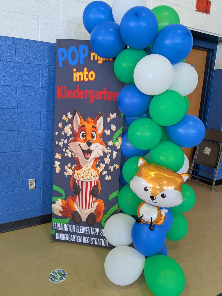 A colorful display in a school hallway features a tall sign that reads “Pop right into Kindergarten!” with an illustration of a smiling cartoon fox holding a bucket of popcorn. Next to the sign is a balloon arrangement in blue, green, and white, including a fox-shaped balloon near the bottom. The display promotes kindergarten registration at Farmington Elementary School.