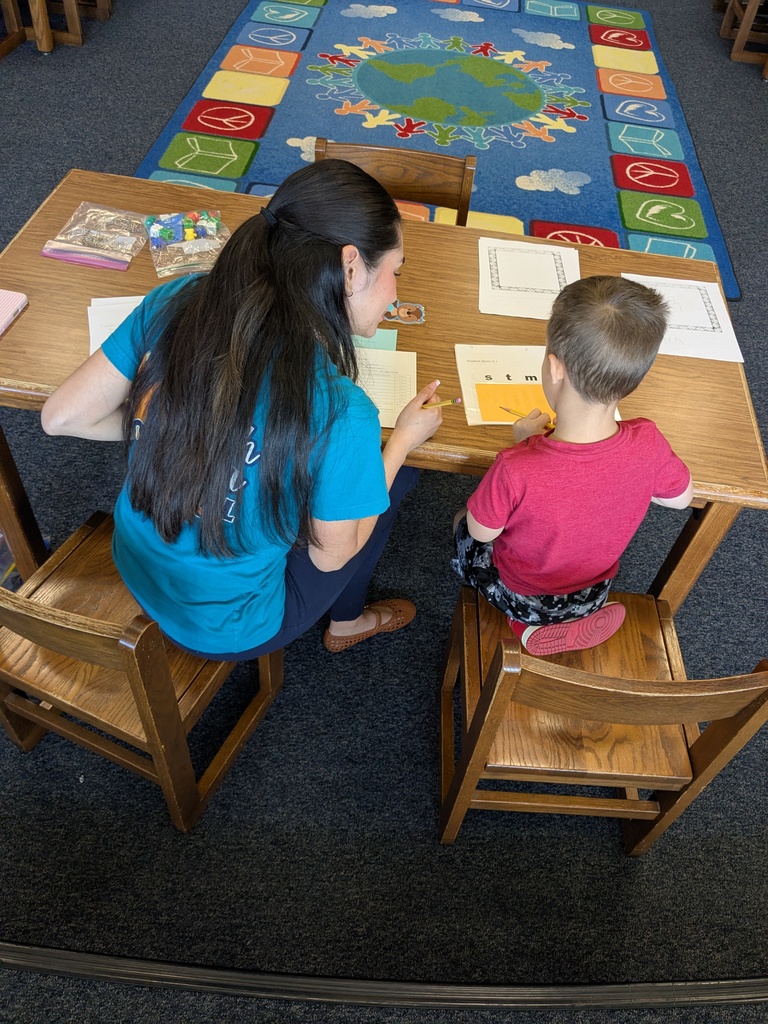 An adult teacher sits beside a young child at a wooden table, both looking down at a worksheet with letters. The teacher points to the paper while the child holds a pencil. A colorful classroom rug with a globe and children holding hands is visible in the background, along with learning materials on the table.