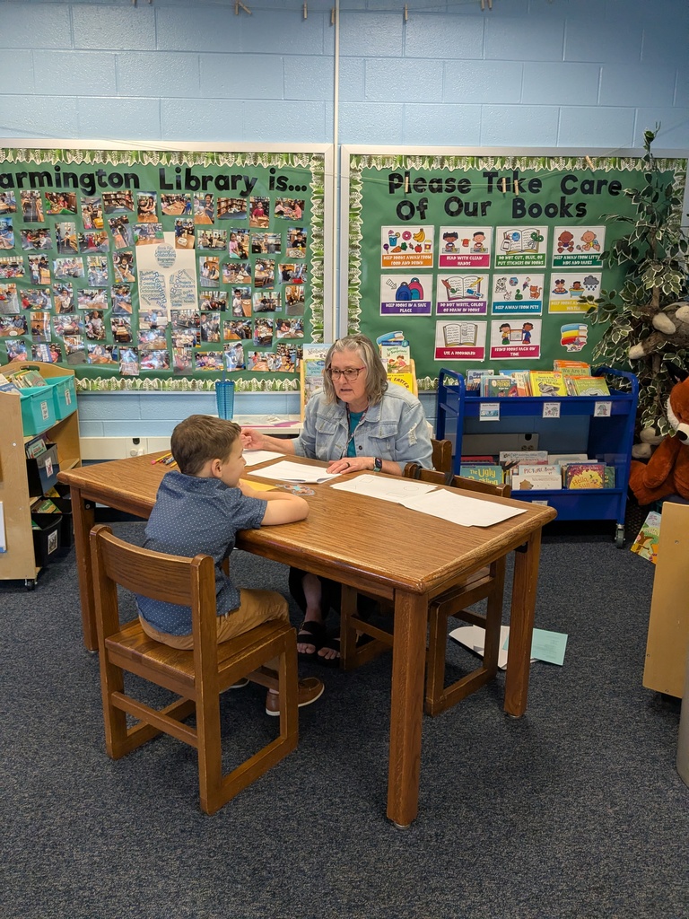 An adult teacher sits at a small wooden table in a school library, working one-on-one with a young child who is seated across from her. Papers and pencils are spread on the table as they focus on a learning activity. Behind them, bulletin boards display photos and library rules, and bookshelves and a reading cart are visible.