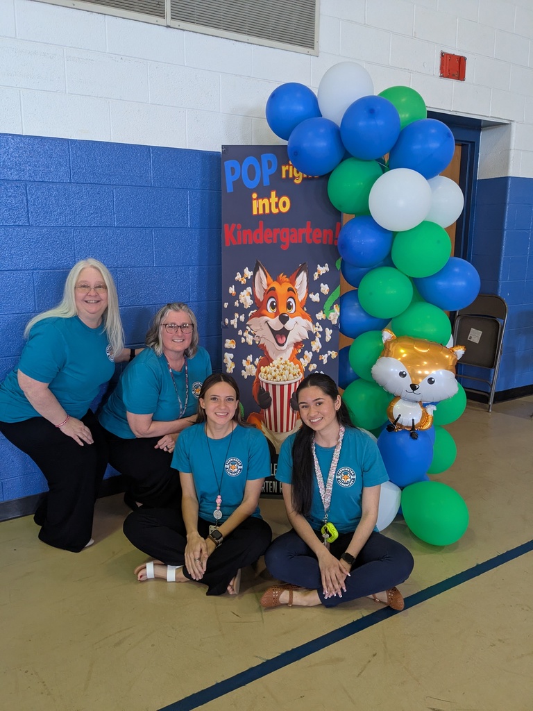Four adults wearing matching shirts pose together in a school gym next to a display with blue, green, and white balloons and a sign that reads “Pop into Kindergarten!” featuring a cartoon fox holding popcorn. Two people kneel in front while two stand behind, smiling at the camera.